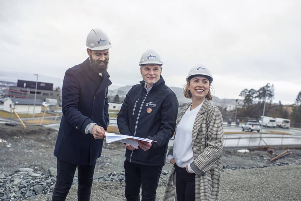 Tre personer på befaring på byggeplass. Hjelmer med NorgesHus på. Studerer plantegning og ser mot noe utenfor kamera.