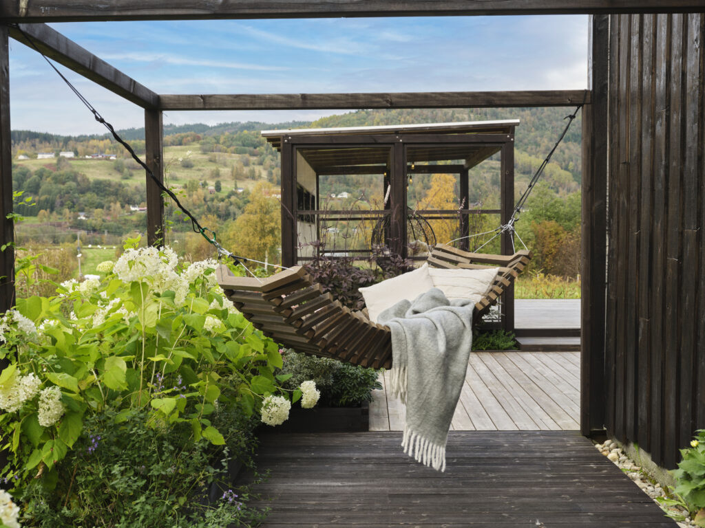 Foto fra Marte Helgetun sin hage, Overbygd pergola i MøreRoyal svart i bakgrunnen. Hengekøye i MøreRoyal grå i forkant. I hengekøyen ligger det myke puter og pledd. Frodige blomster og planter på venstre side av hengekøyen.