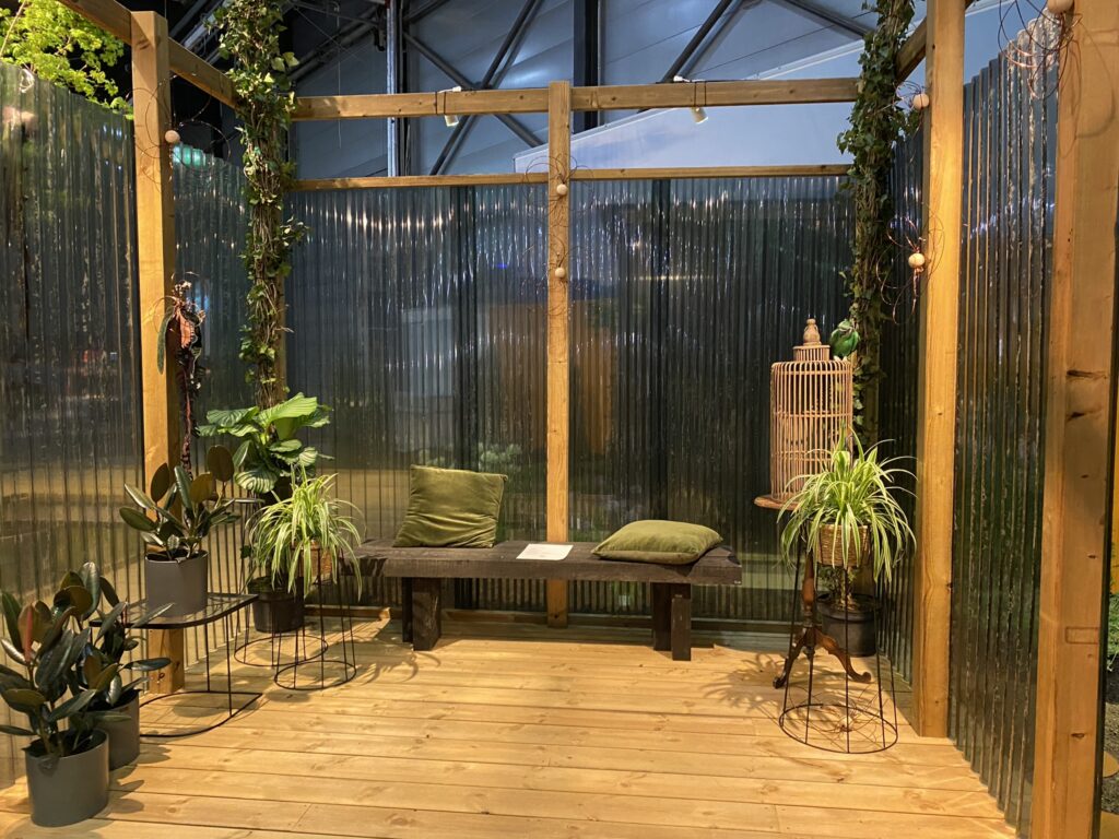 Foto av Fantasihagen fra Hagemessen 2024 - designet av Kjersti Bergesen. Nærbilde av pergola bygd med MøreRoyal og Plastmo. Uten tak. Farge natur. Boardwalk eller promenade i MøreRoyal terrassebord, Concise 28x120. En benk til å sitte på er plassert inne i pergolaen, med to grønne puter. Ulike planter og et fuglebur i tre som blikkfang.